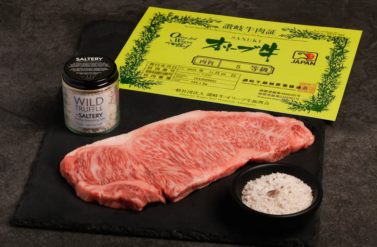 A raw Olive Wagyu strip steak on black slate beside its yellow Japanese grading certificate