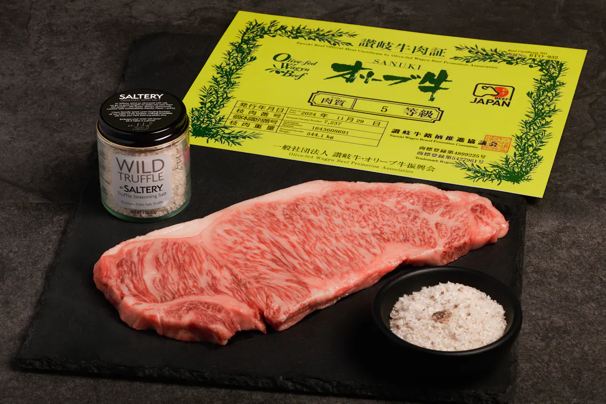 A raw Olive Wagyu strip steak on black slate beside its yellow Japanese grading certificate