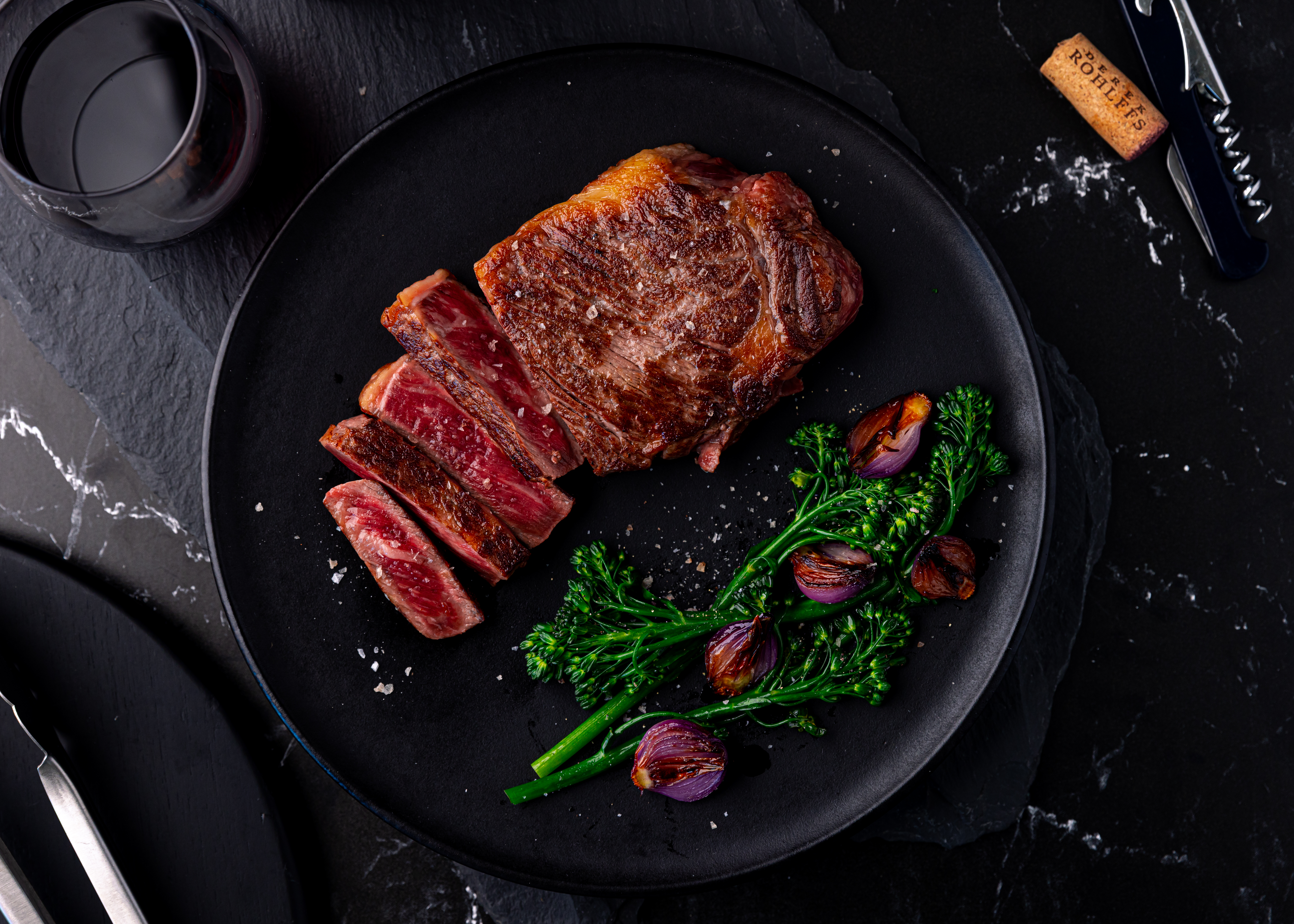 Sliced seared New York strip with broccolini and charred shallots on a black plate, beside a glass of red wine