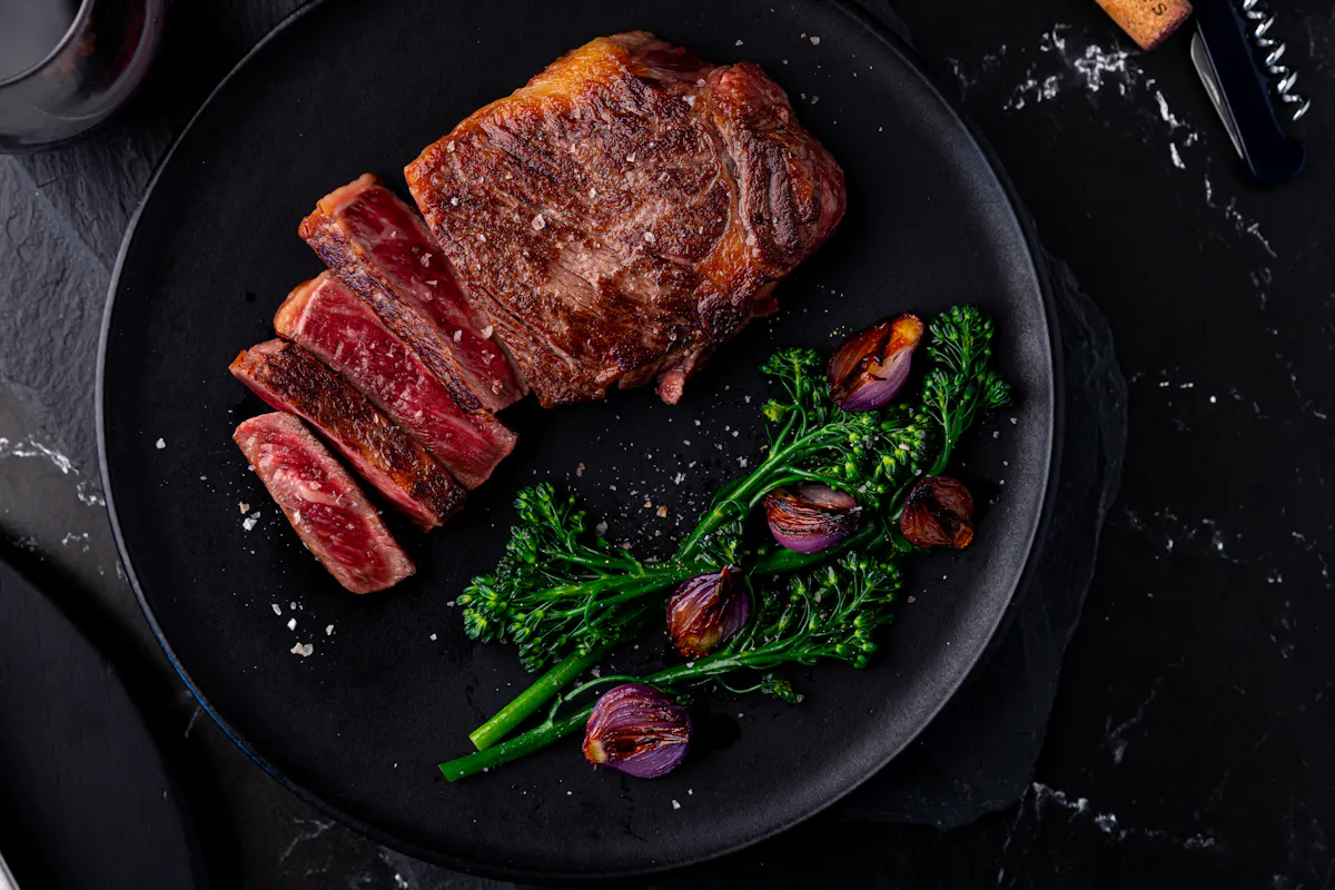 A seared New York strip, partly sliced, on a black plate with broccolini