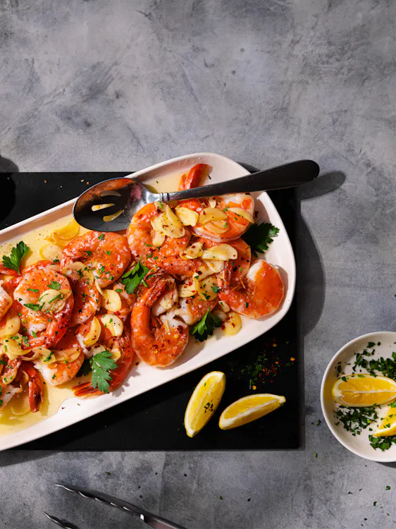 A white platter of cooked jumbo shrimp in garlic butter with herbs and lemon wedges