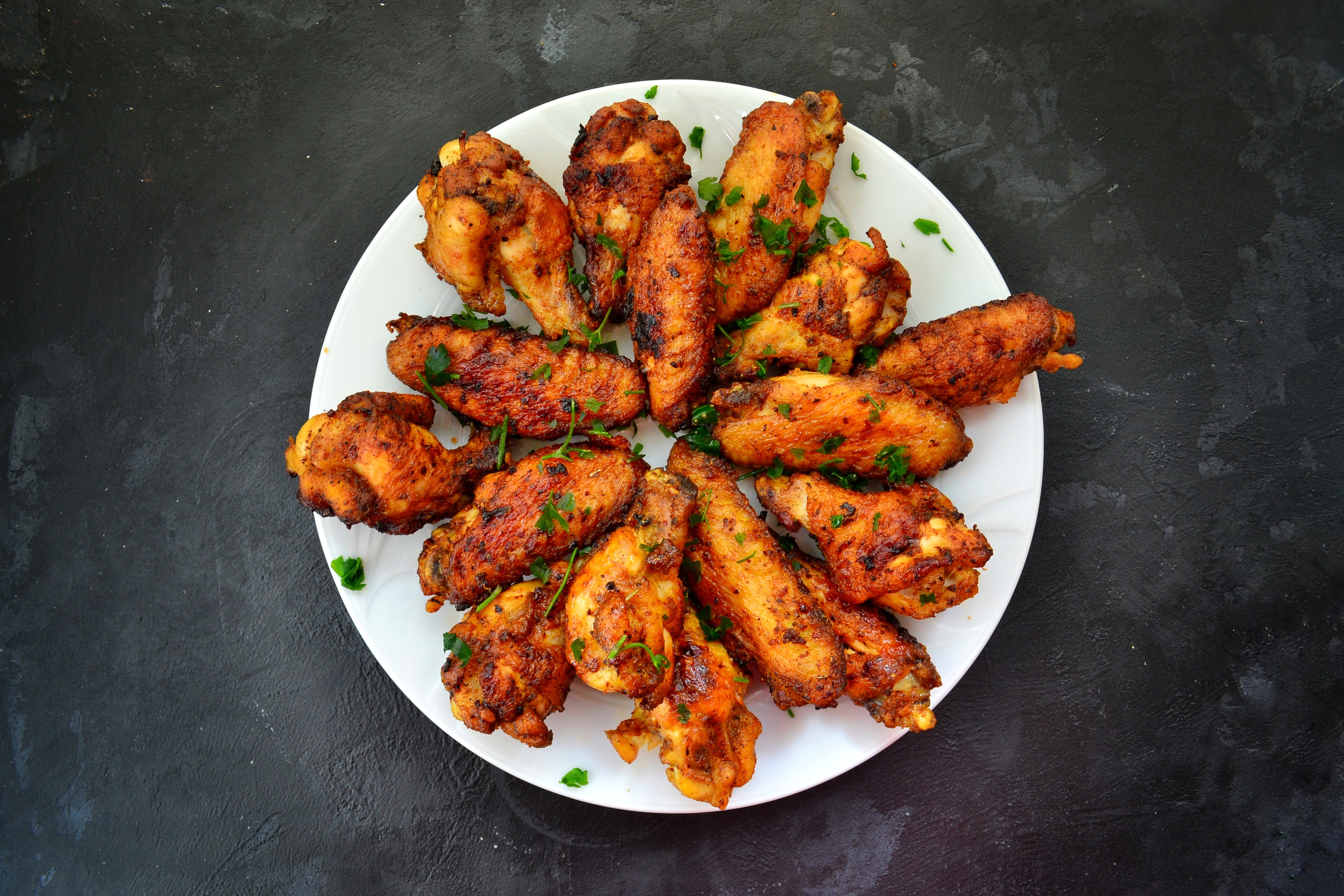 A white plate of seasoned roasted chicken wings with chopped herbs on dark stone