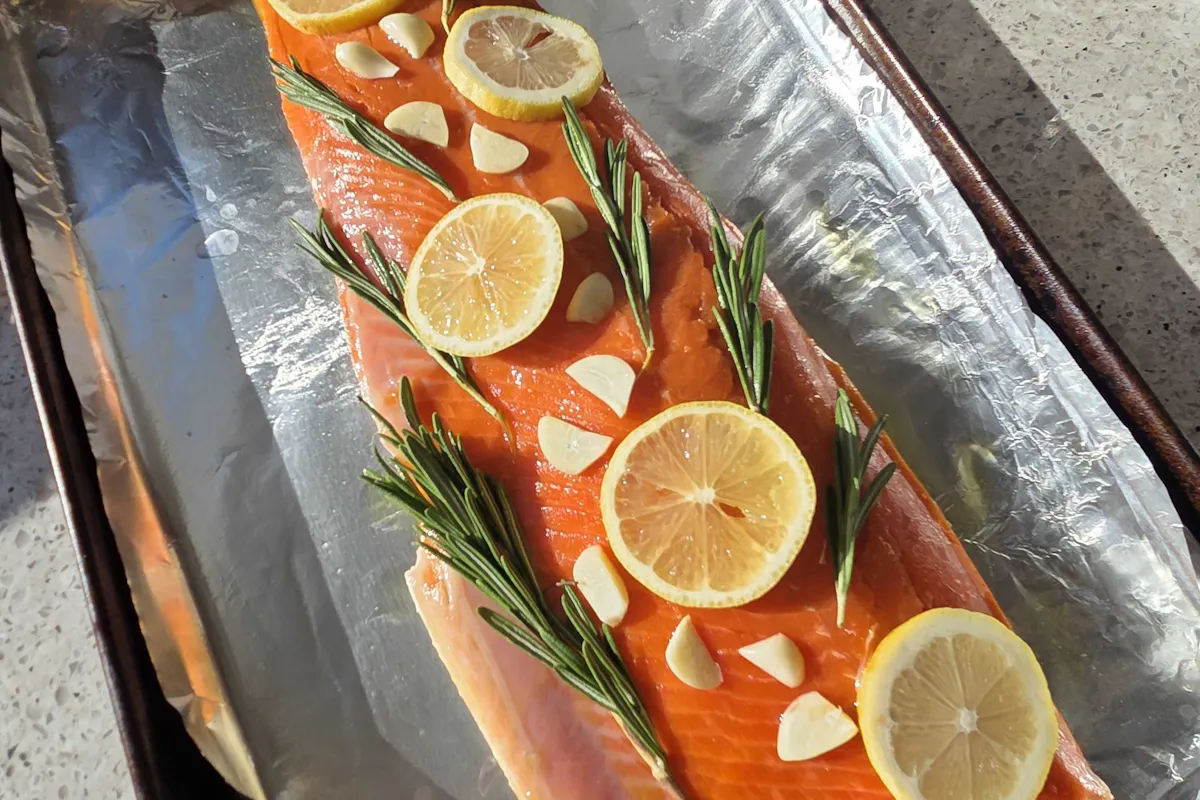 A whole side of Copper River sockeye salmon topped with lemon rounds, rosemary and garlic on a foil-lined tray