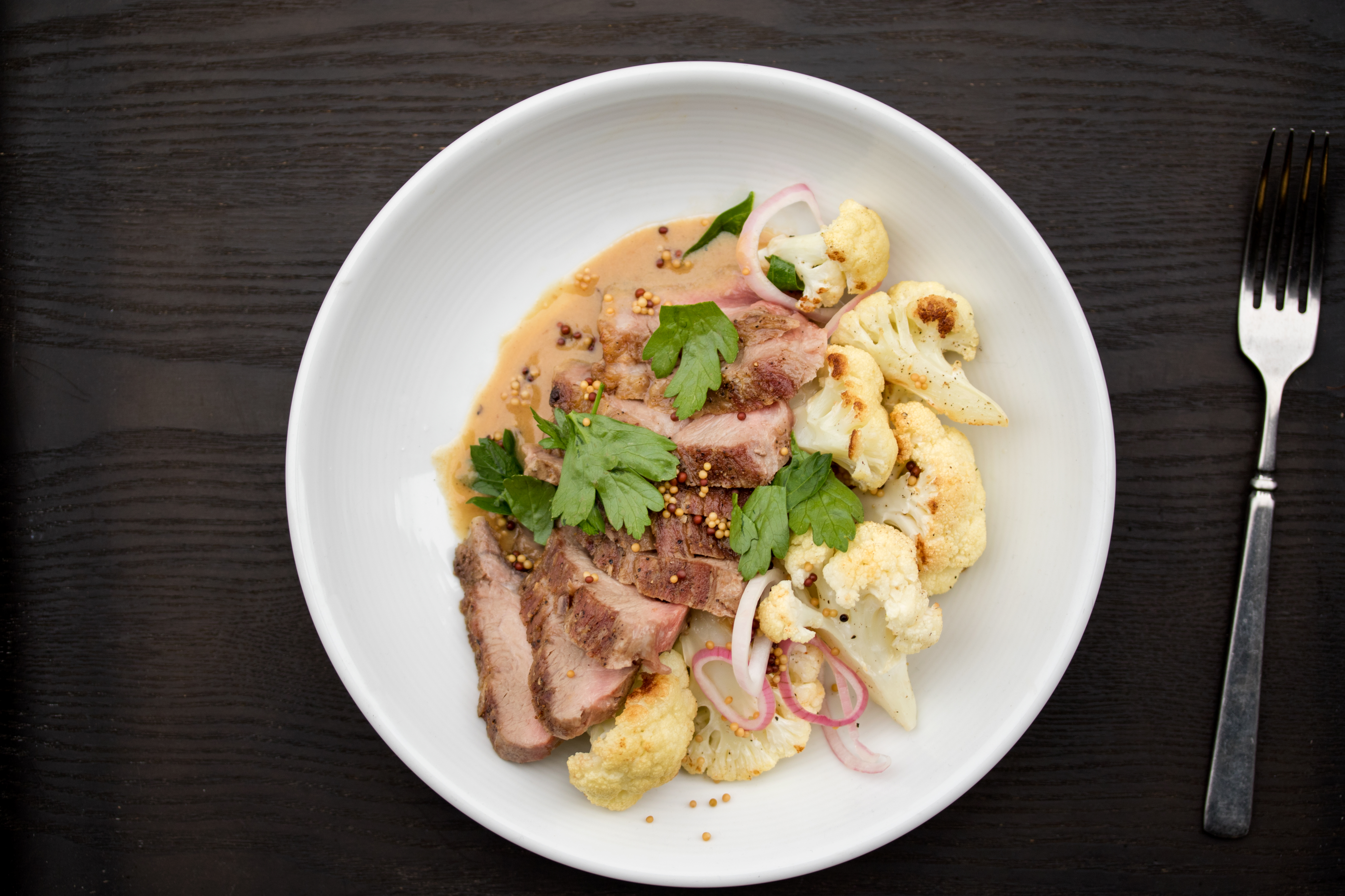Sliced Ib&eacute;rico secreto with roasted cauliflower, pickled red onion and white wine mustard sauce in a white bowl