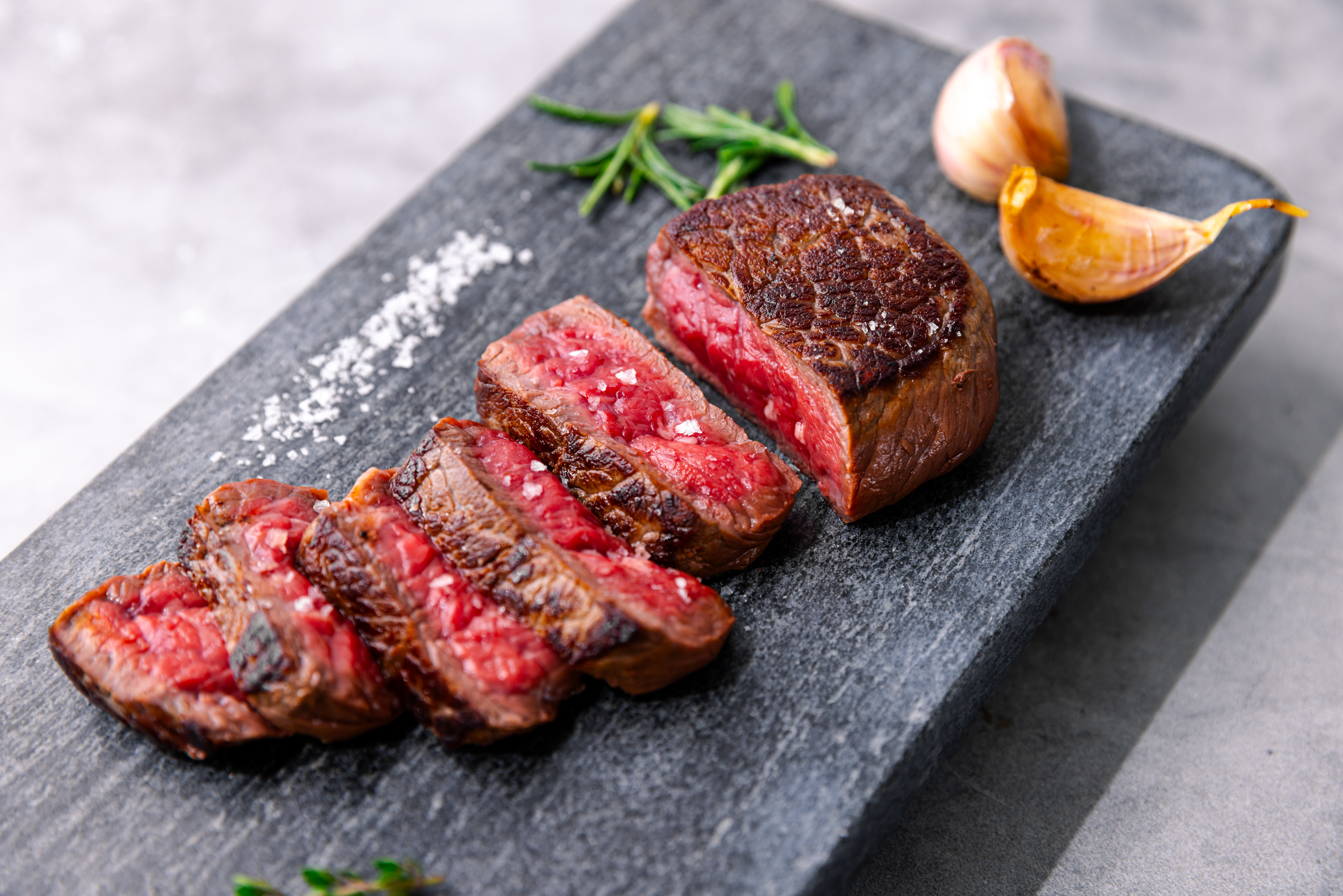 Sliced medium-rare Wagyu top sirloin on a dark stone board with coarse salt, rosemary and roasted garlic