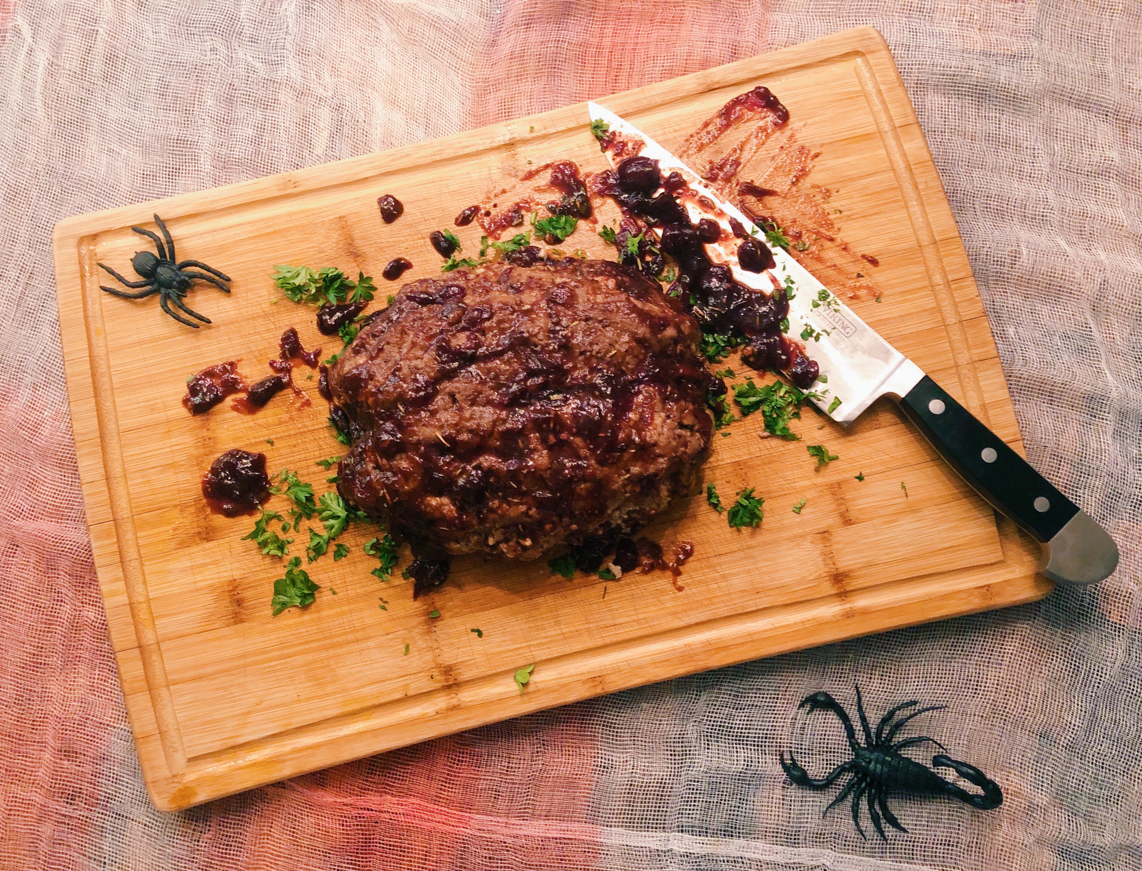 Brain-shaped meatloaf on a wooden board with balsamic cranberry sauce and plastic spiders