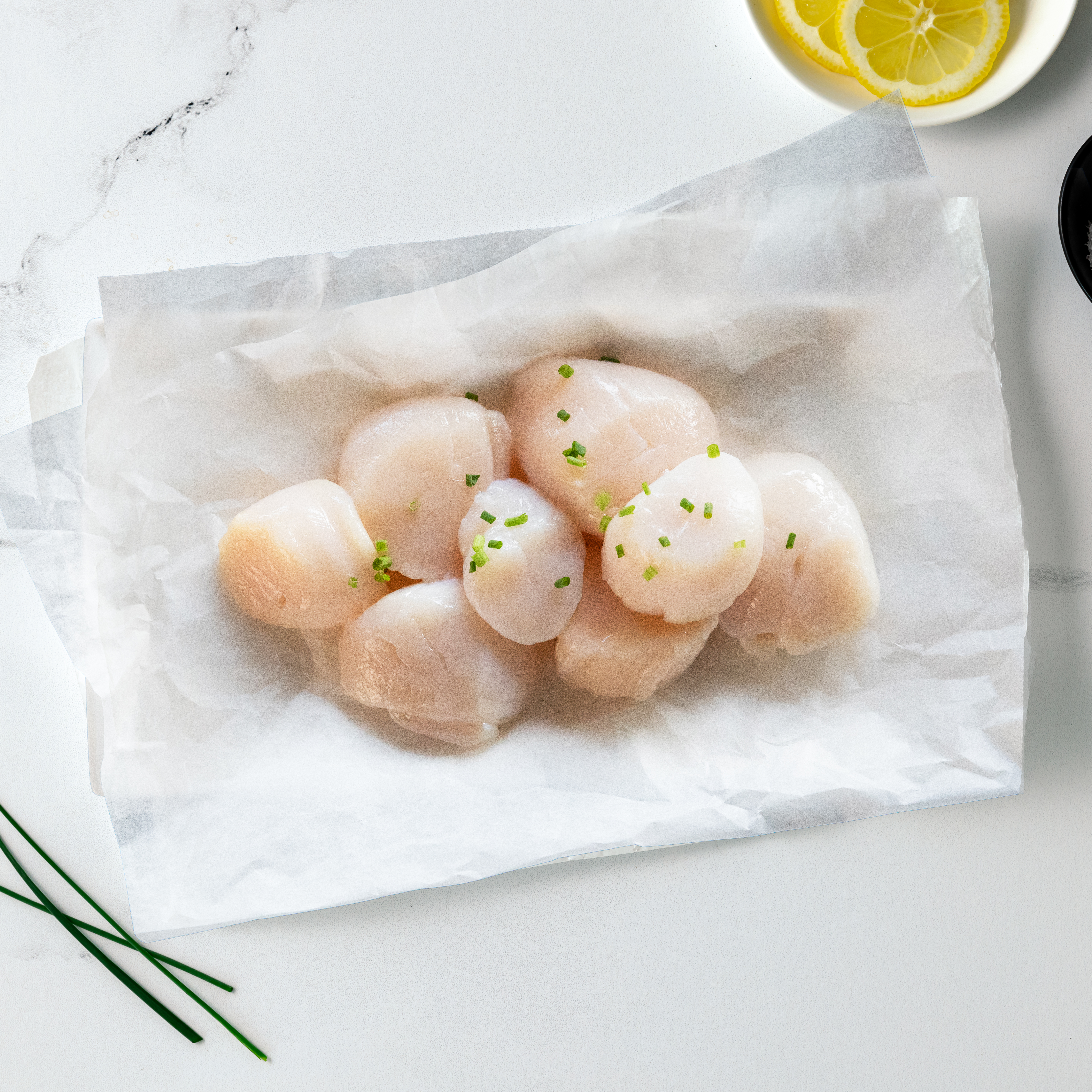Raw jumbo sea scallops piled on parchment paper with lemon slices and chives
