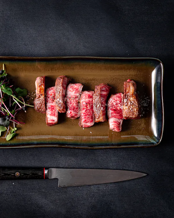Sliced, seared A5 Wagyu petite ribeye on a glazed rectangular platter with microgreens, a chef&rsquo;s knife below