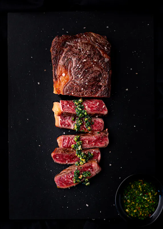 A seared, deeply marbled Wagyu ribeye sliced on a black board with herb sauce