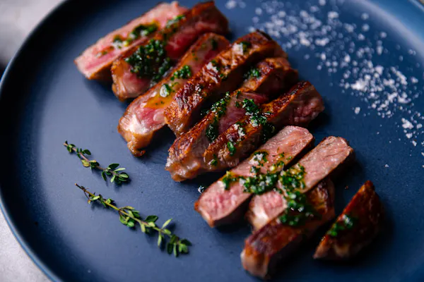 Sliced medium-rare New York strip steak with herb sauce and thyme on a dark blue plate