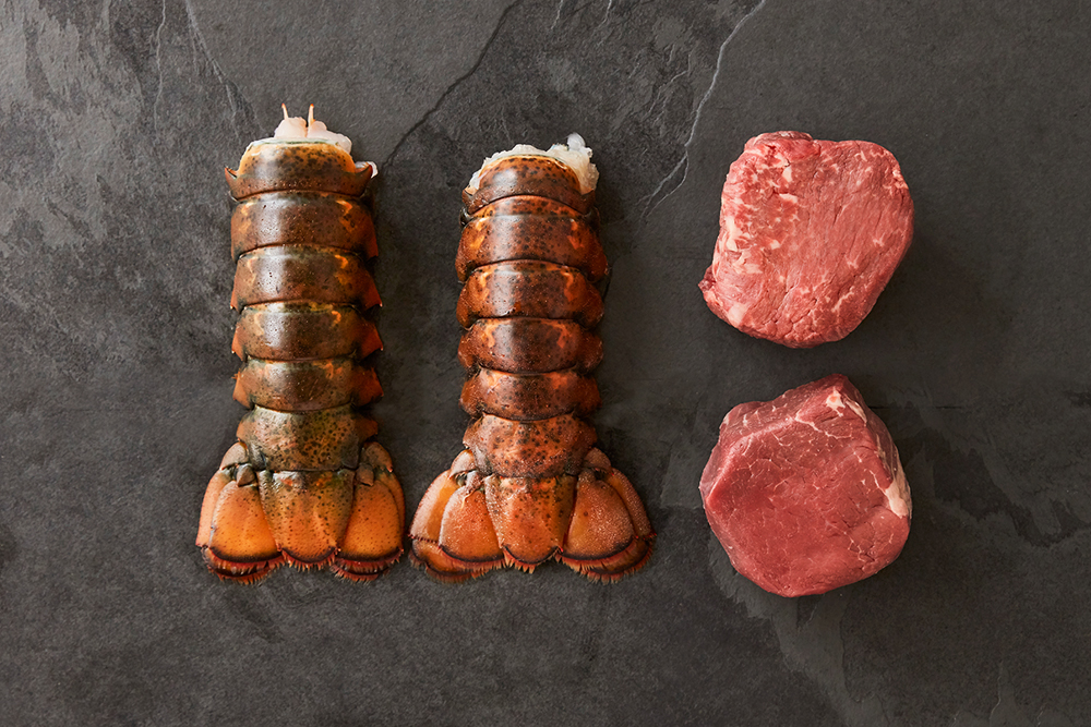 Two raw lobster tails beside two beef tenderloin medallions on dark stone