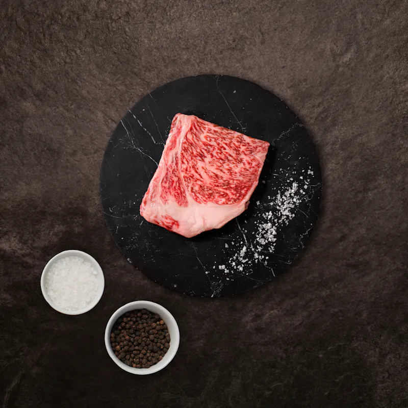 A raw, heavily marbled A5 Wagyu petite striploin on a black plate with salt and pepper