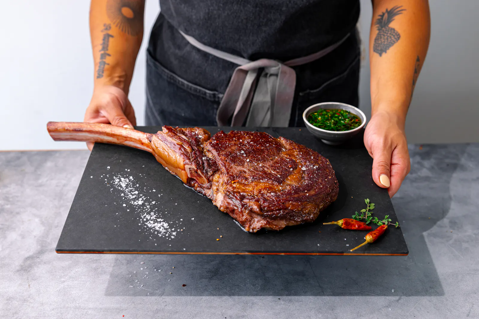A seared Wagyu tomahawk steak with a long frenched bone, served on a black board with flaky salt and chimichurri