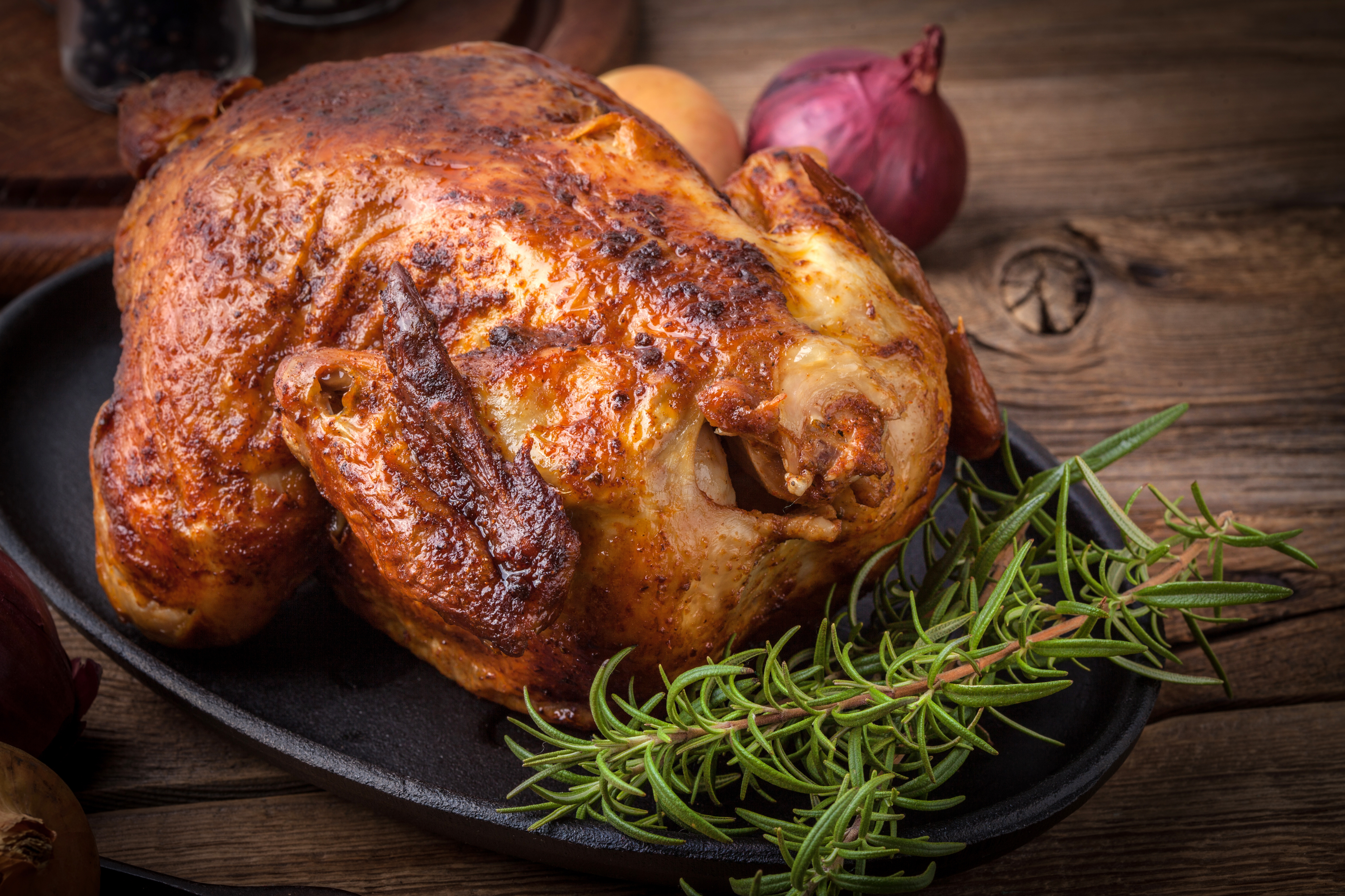 A whole golden roast chicken on a black cast-iron oval with rosemary and red onion