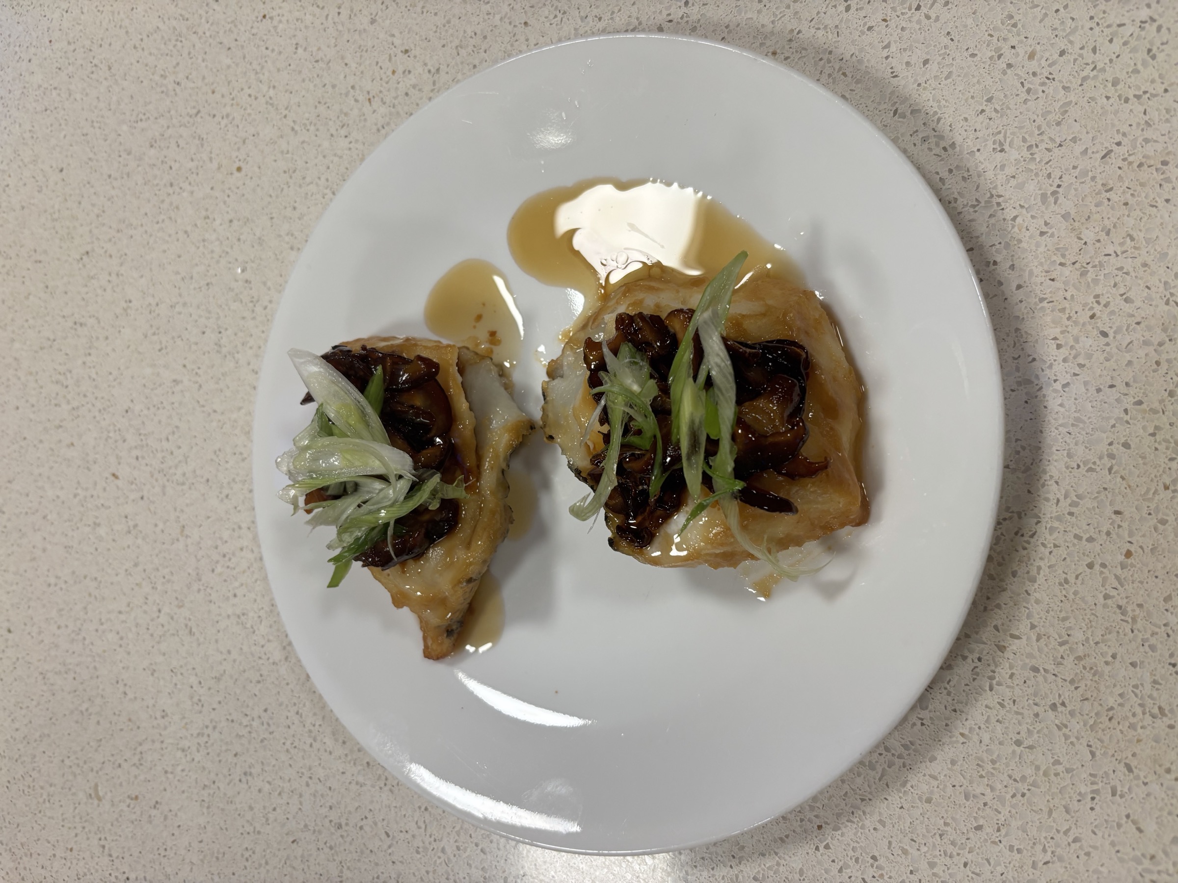 Two pieces of miso-glazed Chilean sea bass topped with shiitake mushrooms and sliced scallions on a white plate