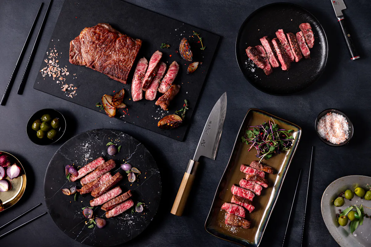 Plates of sliced A5 Wagyu steaks on a dark table with olives, salt and a chef’s knife