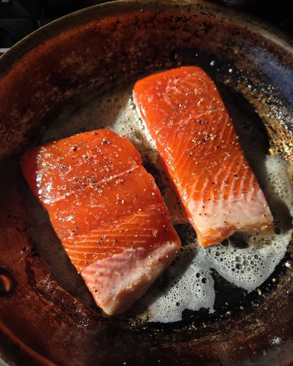 Two Copper River salmon fillets searing flesh-side up in a stainless steel skillet, glistening orange with cracked pepper
