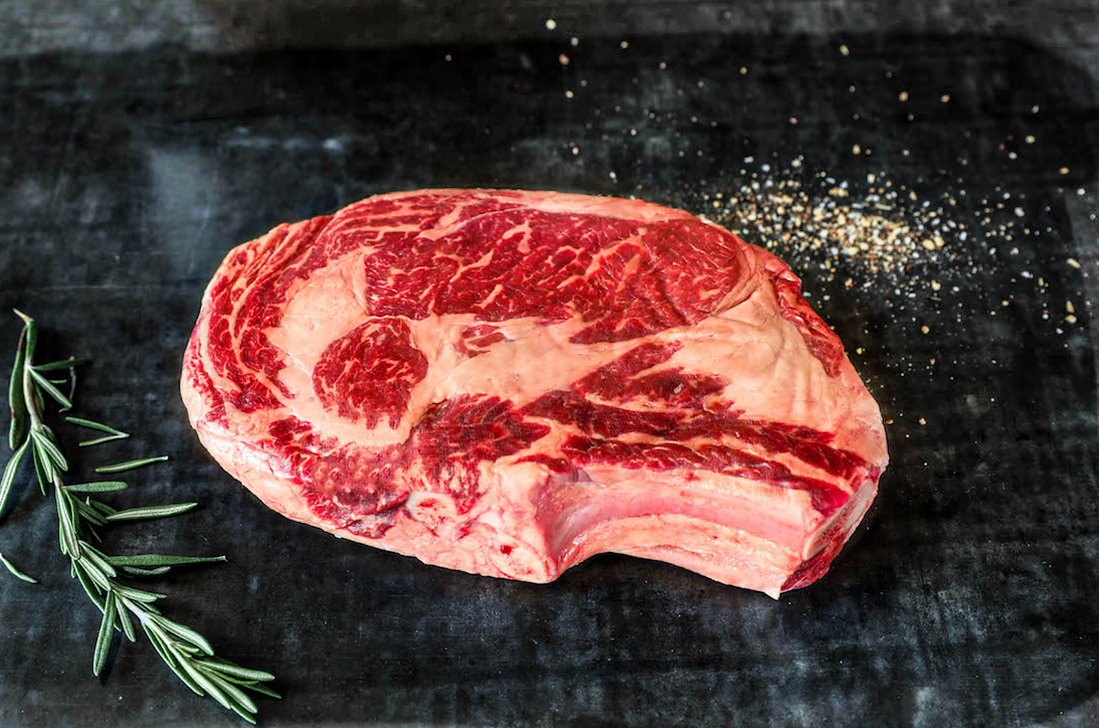 A raw bone-in Wagyu-cross ribeye steak with heavy marbling, with rosemary and seasoning
