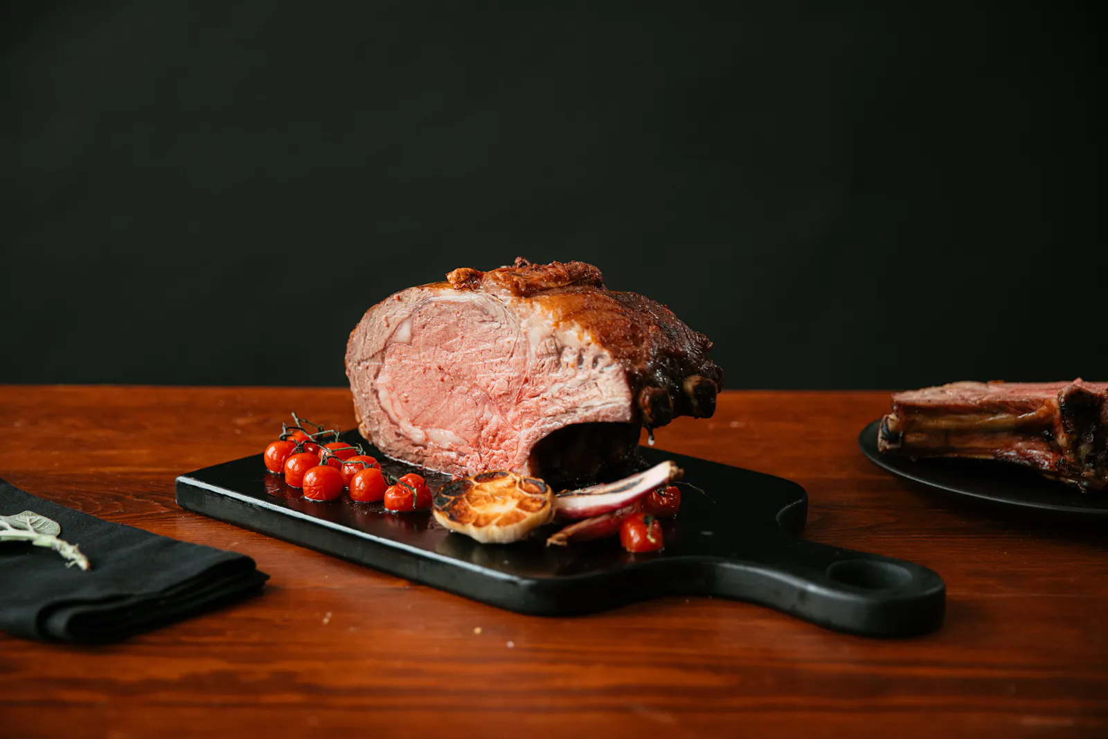 A bone-in prime rib roast, sliced to show its rosy center, on a black serving board with roasted tomatoes and garlic