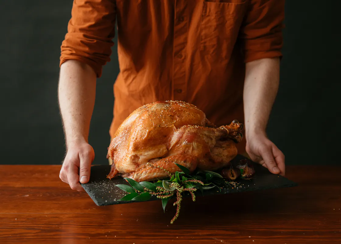 A person carries a whole roasted turkey on a slate board garnished with greenery