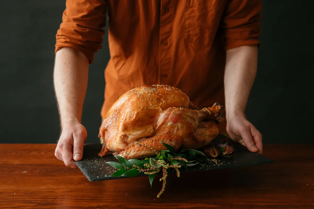 A person carries a whole roasted turkey on a slate board garnished with greenery