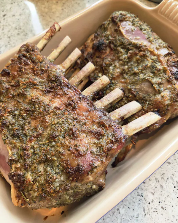 Two herb-crusted frenched lamb racks roasted golden in a cream baking dish, bones crossed